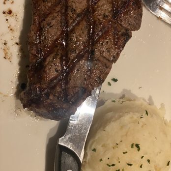Bonefish Grill - Filet Mignon* with potatoes - A cooked filet mignon steak with visible grill marks is placed on a white plate, accompanied by a serving of mashed potatoes topped with chopped herbs. A fork and a steak knife are also visible on the plate, indicating the meal is ready to be eaten. The presentation suggests a prepared dish served at a restaurant or home setting. - Seafood Near Me - Gambrills, Maryland