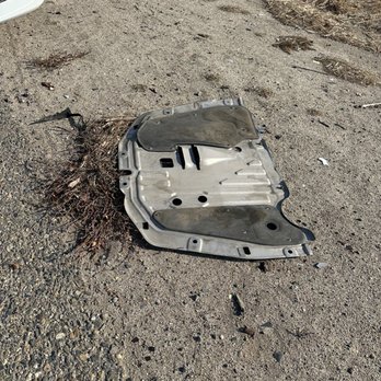 Honda North - Oil pan guard - A metal oil pan guard is positioned on a gravel and dirt surface outdoors, with some dried vegetation around it. The guard appears used, with dirt and wear visible. A portion of a white vehicle's front bumper and headlight is seen in the top left corner of the image, indicating the location is near a vehicle, likely at a repair or maintenance site. - Auto Repair Near Me - Clovis, California