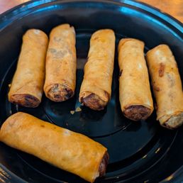 Spring Rolls - Six spring rolls are arranged in a black tray, with one partially unwrapped showing dark filling inside.