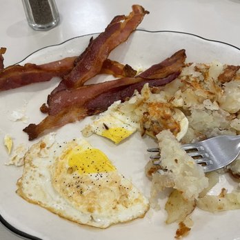 Peppercorn - 2 Eggs bacon and home fries - The image shows a breakfast plate with several strips of crispy bacon, a fried egg with a runny yolk, and a portion of home fries. The plate is white with a black rim, and the setting includes a salt shaker, a spoon, and a small container in the background. The meal appears to be served in a casual dining environment. - Diners Near Me - Summit, New Jersey