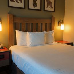 Room - The image shows a hotel room with a neatly made bed that has white linens and three pillows. The bed has a wooden headboard, and above it, there are three framed pictures of animals with plaid or patterned silhouettes: a bear, a deer, and a wolf, arranged in a row on a green wall. On each side of the bed, there are wall-mounted lamps providing warm lighting. Small bedside tables are visible, with one holding a telephone and a clock. The room has a cozy, rustic decor style.