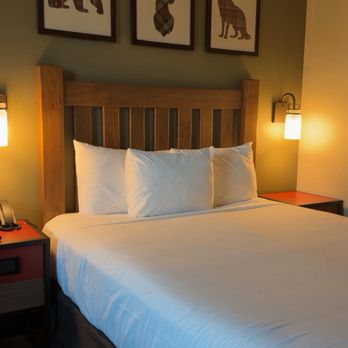 Great Wolf Lodge - Room - The image shows a hotel room with a neatly made bed that has white linens and three pillows. The bed has a wooden headboard, and above it, there are three framed pictures of animals with plaid or patterned silhouettes: a bear, a deer, and a wolf, arranged in a row on a green wall. On each side of the bed, there are wall-mounted lamps providing warm lighting. Small bedside tables are visible, with one holding a telephone and a clock. The room has a cozy, rustic decor style. - Resorts Near Me - Mashantucket, Connecticut