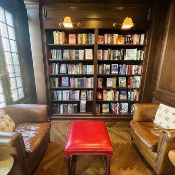 Flyleaf - Private spaces - The image shows a cozy reading area with two brown leather armchairs, each with a decorative pillow, positioned near large windows with grid panes. Between the chairs is a red upholstered ottoman. Behind the chairs is a tall, dark wooden bookshelf filled with a variety of colorful books. The space has wooden paneling on the walls and warm lighting from two wall-mounted lamps, creating an inviting atmosphere for reading or relaxing. - Bookstores Near Me - Grosse Pointe Farms, Michigan