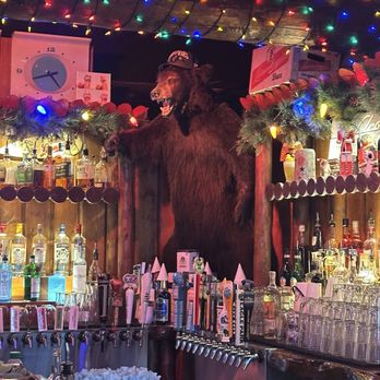 Camp Bar Third Ward - Bar - A mounted bear head with an open mouth is displayed behind a bar area, adorned with Christmas garlands and multicolored string lights. The bar features various bottles of alcohol, beer taps, and glassware, with festive decorations including small figurines and holiday-themed items. A clock is visible on the wall above the display, and the overall setting suggests a rustic, holiday-themed bar environment. - Bars Near Me - Milwaukee, Wisconsin