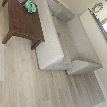 Floor Daddy - A beige fabric sectional sofa with multiple cushions is placed in a living room with light wood flooring. A dark wooden coffee table is positioned in front of the sofa, holding various items including a green cloth, a small container, and some tools. The room features a staircase with a wooden railing, a mirror on the wall, and a partial view of a kitchen or dining area in the background. - Flooring Near Me - Tempe, Arizona