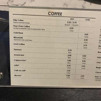 Usagi NY - Coffee Menu as of November 2021 - The image shows a coffee menu with a list of beverages including drip coffee, pour-over, cold brew, mizudashi, iced coffee, espresso, americano, cappuccino, latte, café au lait, and mocha. Prices are listed for both hot and iced options, with some items marked with an asterisk. The menu is printed on white paper and placed inside a clear display frame on a dark surface, with a marble countertop visible in the background. - Bookstores Near Me - Dumbo, New York