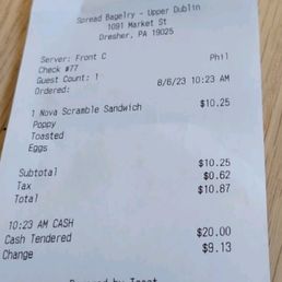 A paper receipt from Spread Bagelry located at 1091 Market St, Dresher, PA, is shown on a wooden surface. It details a purchase of a Nova Scramble Sandwich with toasted eggs, totaling $10.87, paid in cash. The receipt includes the date and time of the order, server information, and payment details.