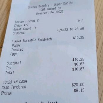 Spread Bagelry - A paper receipt from Spread Bagelry located at 1091 Market St, Dresher, PA, is shown on a wooden surface. It details a purchase of a Nova Scramble Sandwich with toasted eggs, totaling $10.87, paid in cash. The receipt includes the date and time of the order, server information, and payment details. - Bagels Near Me - Dresher, Pennsylvania
