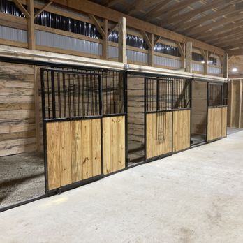 Blackstone Stables - Boarding Stalls - The image shows a row of horse stalls constructed with wooden panels and black metal bars, situated inside a large indoor equestrian facility. The stalls are aligned along a concrete floor, with a wooden upper structure and ceiling. The background includes additional wooden walls and lighting fixtures, indicating a spacious stable designed for horse boarding and care. - Horseback Riding Near Me - Howell, Michigan
