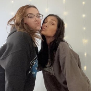 Sweets On Main - Us at their selfie wall pretty plain tho - Two women are posing together in front of a wall decorated with vertical strings of small, warm-colored lights. They are leaning toward each other, with puckered lips as if about to kiss. One woman has light brown hair, glasses, and is wearing a dark hoodie, while the other has dark hair and is wearing a gray jacket. The setting appears to be indoors, with a casual and friendly atmosphere. - Ice Cream & Frozen Yogurt Near Me - Fort Wayne, Indiana