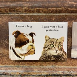 A greeting card displays a dog and a cat with captions about hugs, set against a wooden background.