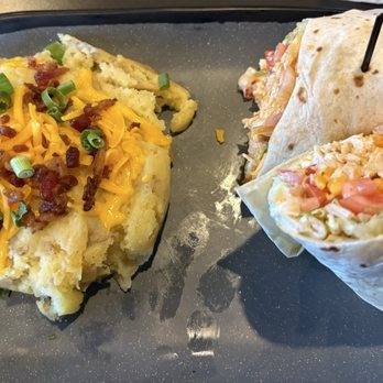 TJ Chumps - Buffalo Chicken wrap & loaded mashed potatoes - A black tray holds two wraps, each secured with a black toothpick, filled with shredded lettuce, tomatoes, and possibly chicken or other ingredients. Next to the wraps is a serving of mashed potatoes topped with shredded cheddar cheese, bacon bits, and chopped green onions. The presentation suggests a casual meal, likely from a restaurant or takeout service. - Chicken Wings Near Me - Kettering, Ohio