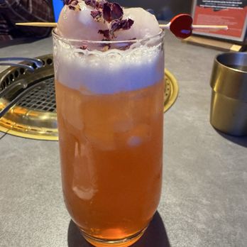 Y Space BBQ - A tall, clear glass holds a pinkish-orange cocktail with a foamy top, garnished with dried flower petals and a cherry on a skewer. The drink is placed on a restaurant table with a grill and a metal cup nearby, and a menu or sign is visible in the background. The setting appears to be a restaurant or bar with a modern interior. - Barbeque Near Me - Flushing, New York