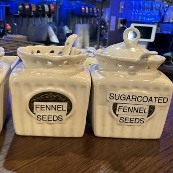 Monsoon Grill - Ceramic spice jars labeled for fennel seeds and sugarcoated fennel seeds are placed on a bar or restaurant counter with bottles and bar equipment in the background. - Indian Near Me - Topeka, Kansas