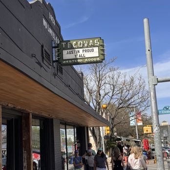 Tecovas - A busy street scene in Austin, Texas, shows pedestrians walking along the sidewalk in front of a storefront with large windows and a black exterior. A sign above the store reads "Tecovas" with additional text "Austin Proud" and "Western Goods." The sky is clear with some clouds, and there are trees and street signs visible, including a street sign for West Elizabeth Street. The scene captures a typical urban environment with people casually strolling and shopping. - Leather Goods Near Me - Austin, Texas
