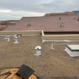1--Beginning roof removal - The photograph captures a rooftop with several vents and a skylight installed on a flat surface. The vents are white and metallic, positioned at different points across the roof. The skylight has a rectangular shape with a dark frame. The background features a cloudy sky and distant mountains. In the foreground, roofing materials and tools are visible, indicating ongoing roofing work or maintenance.