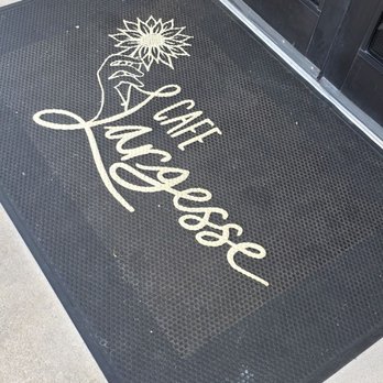Cafe Largesse - A black rubber doormat featuring the words 'Cuff Luvesse' in cursive script and a stylized flower illustration, positioned in front of a glass door with a metal frame. The doormat is on a concrete surface near a stone wall, indicating an entrance to a business or establishment. - Cafes Near Me - Sunset Valley, Texas