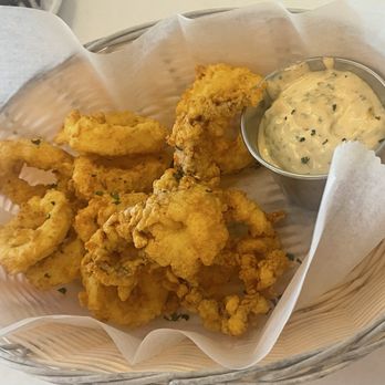 Oceanview Diner - Calamari  Soo good - The image shows a basket containing fried calamari with a golden, crispy coating. The calamari pieces are arranged on a paper lining inside the basket. A small metal cup filled with a creamy, herb-infused dipping sauce is placed next to the calamari. The basket is lined with parchment paper, and the setting suggests a casual dining environment, likely at a diner or seafood restaurant. - Breakfast & Brunch Near Me - Norfolk, Virginia