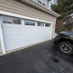 The image shows a beige house with horizontal siding and a white sectional garage door featuring four small windows at the top. The garage door is closed, and a black truck with a V8 badge is parked on the asphalt driveway in front of it. A small exterior light is mounted on the house near the garage. The background includes trees and a partly cloudy sky.