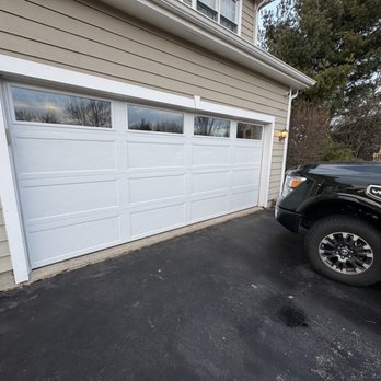 Anytime Garage Door - The image shows a beige house with horizontal siding and a white sectional garage door featuring four small windows at the top. The garage door is closed, and a black truck with a V8 badge is parked on the asphalt driveway in front of it. A small exterior light is mounted on the house near the garage. The background includes trees and a partly cloudy sky. - Garage Door Services Near Me