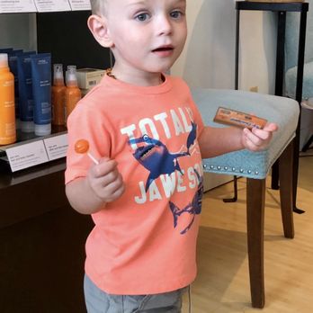 Panache - A boy with blond hair and blue eyes, wearing a coral T-shirt with a shark graphic, holds a lollipop and a card inside a retail store. - Hair Salons Near Me - St. Augustine, Florida