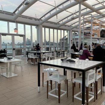 Offshore Rooftop - A rooftop restaurant with large glass windows offering views of the city skyline, featuring white and black tables, chairs, and a bar area with patrons. - New American Near Me - Chicago, Illinois