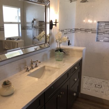 Modesto Kitchen & Bath - Bathroom - The image shows a bathroom with a marble countertop vanity, a large framed mirror, and a flower arrangement in a glass vase. The vanity has dark cabinetry with silver knobs. The shower area has a wall-mounted showerhead, a small window, and a decorative glass tile accent wall. The bathroom has neutral tones and a wooden floor mat. - Interior Design Near Me - Modesto, California