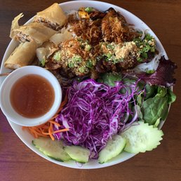 Bamboo Vermicelli Noodle Bowl with Grill Angus Beef Skewer, shrimp, and egg roll - A Vietnamese restaurant dish presented on a white plate includes spring rolls, grilled meat topped with chopped green onions and crushed peanuts, shredded purple cabbage, cucumber slices, and mixed greens. A small bowl of dipping sauce is placed on the side. The dish is served on a wooden table, showcasing a variety of fresh ingredients and traditional flavors.