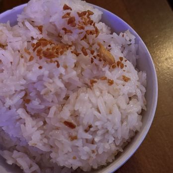 FOB Kitchen - Garlic rice - A close-up image of a bowl filled with cooked white rice, topped with small pieces of fried garlic. The rice appears fluffy and well-cooked, with the garlic adding a crispy texture. The bowl is placed on a wooden surface, suggesting a dining environment, possibly at a Filipino restaurant. - Filipino Near Me - Oakland, California