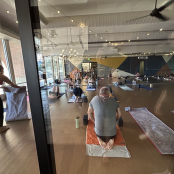 The Foundry - Yoga room - The image shows a yoga class inside a studio with large windows allowing natural light, string lights hanging from the ceiling, and a wooden floor. Several participants are on yoga mats, some in kneeling or seated positions, with a person at the front assisting a participant. The studio has a modern, open design with a colorful geometric wall in the background. - Yoga Near Me - Mesa, Arizona
