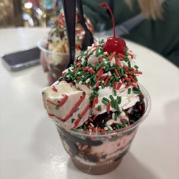 Jingle all the way (december special) - A dessert cup filled with whipped cream, topped with colorful sprinkles, a cherry, and a marshmallow, served in a clear plastic cup.