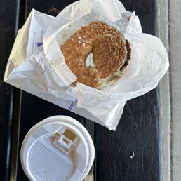 A partially eaten bagel with cream cheese is wrapped in white paper on a black bench, accompanied by a disposable coffee cup with a lid.