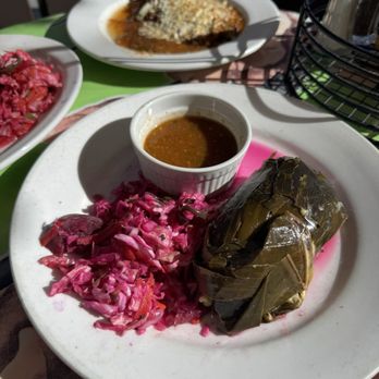 Tune-Up Cafe - Banana leaf wrapped Chicken tamale. - A white plate features a banana leaf-wrapped tamale, accompanied by pink pickled cabbage and a small cup of sauce. In the background, there are additional dishes, including a plate with a dish topped with cheese and sauce. The setting appears to be outdoors with sunlight casting shadows, and a basket with condiments is also visible. - New Mexican Cuisine Near Me - Santa Fe, New Mexico