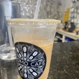 Hazelnut Horchata Latte - A clear plastic cup with a lid and straw contains a hazelnut horchata latte from Oromo Cafe. The cup has condensation on the outside, indicating a cold beverage. It is placed on a dark, speckled countertop, with a water bottle and cafe interior visible in the background, including shelves and equipment.