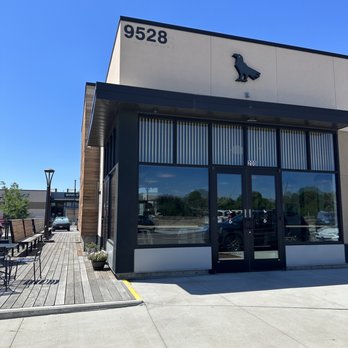 Corvus Coffee Roasters - Door is on the back side of the building. Plenty of parking. - A commercial building with large glass windows, the number 9528, and a dinosaur logo on the facade, with outdoor seating and parking. - Coffee & Tea Near Me - Arvada, Colorado