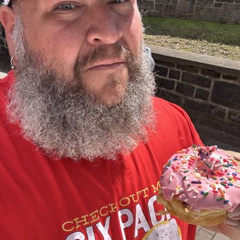 Donerds Donuts - The Homer - A man with a gray beard, wearing a red shirt and a cap, holds a pink frosted donut with colorful sprinkles. - Donuts Near Me - Bethlehem, Pennsylvania