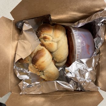 Pegasus Pizza - Gnots - A brown paper takeout box holds a bread roll, a cup of iced coffee, and a small container of sauce, with foil lining inside. - Pizza Near Me - Eugene, Oregon