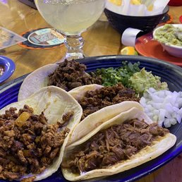 Carne Asada - A plate of four tacos filled with seasoned meat, topped with chopped onions and cilantro, served with lime wedges. A margarita with a straw is in the background, along with condiments and chips.