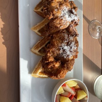 Johnny Pistolas - Chicken & waffles - Three pieces of fried chicken with crispy, golden-brown coating are placed on waffle pieces on a rectangular white plate. The chicken is dusted with powdered sugar. A small bowl of fruit salad containing strawberries, pineapple, and melon is positioned beside the plate. The meal is set on a wooden table with sunlight casting shadows. - Mexican Near Me - Washington, DC, District of Columbia