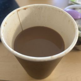 $7.50 Coffee (before tip) before I had taken a sip. - A paper cup filled with a dark brown hot beverage, possibly coffee or tea, is positioned on a light wooden surface. The cup has a small stain or drip on the rim and is surrounded by various objects, including a purple spoon and some packaging, indicating a casual setting.