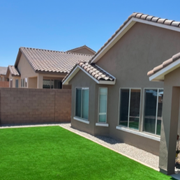 Corner of the house with artificial turf, rock fill in the planter areas, and colored concrete borders. - The backyard features a green artificial turf lawn, gravel areas, and a beige house with large windows and a covered patio.