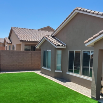 AMG Landscape - Corner of the house with artificial turf, rock fill in the planter areas, and colored concrete borders. - The backyard features a green artificial turf lawn, gravel areas, and a beige house with large windows and a covered patio. - Irrigation Near Me