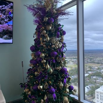 Dauphin's - Mardi Gras! - A Christmas tree is decorated with purple and gold ornaments, ribbons, and string lights. The tree is topped with an arrangement of purple and gold foliage and decorative elements. It is placed indoors near a large window that offers a view of a cityscape with buildings and a cloudy sky. The tree is set on a fabric-covered base, and part of a television screen displaying an underwater scene is visible on the left side of the image. - Seafood Near Me - Mobile, Alabama