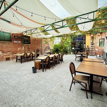 Behind Bars - Quaint courtyard they share with the restaurant next door - A spacious outdoor patio features wooden tables, chairs, string lights, and greenery, enclosed by brick walls and a glass door. - Bars Near Me - Pasadena, California
