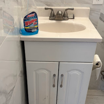 Crossfire Construction - Before, showing the gap that needed a trim peice. See how the top is flush with the wall on the left. - A bathroom vanity features a white cabinet with two doors, a sink with a faucet, and a countertop. A bottle of window cleaner is placed on the left side of the countertop, and a roll of toilet paper is mounted on the wall to the right. The background includes tiled walls and an electrical outlet. - Decks & Railing Near Me