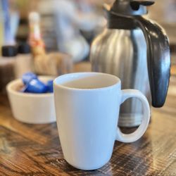 First Watch - Ceramic mug with piping hot coffee carafe in the background.