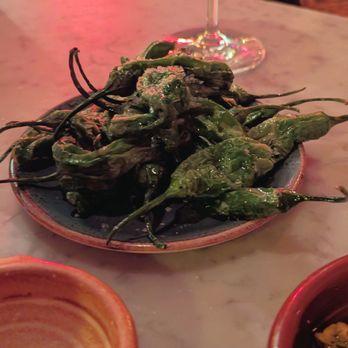 Bar Isabel - A dish of roasted green peppers on a ceramic plate, with a wine glass and small bowls nearby, set on a marble table. - Spanish Near Me - Toronto, Ontario