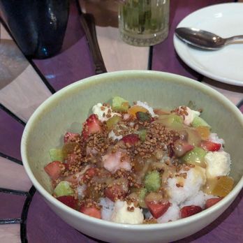 Mariscos San Pedro - Granizado - A bowl of shaved ice dessert features chopped strawberries, green fruit pieces, syrup drizzled on top, and crunchy toppings. It is placed on a table with a white plate, a spoon, and glasses in the background, suggesting a dining setting. - Mexican Near Me - Chicago, Illinois