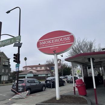 Smokehouse - A round sign for Smokehouse, a restaurant established in 1951, is mounted on a pole near a sidewalk. The sign features a red background with white text, indicating the restaurant's name and offerings such as flame-grilled hamburgers and hot dogs. The surrounding area includes parked cars, a street corner with traffic lights, and nearby residential buildings. The sky is overcast, and there is a small patch of soil with a trash bin nearby. - Fast Food Near Me - Berkeley, California