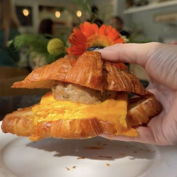 Verzenay - Egg & Cheese on Croissant with Chicken Sausage - A croissant sandwich with scrambled eggs, sausage patty, and melted cheese is held in a person's hand, with a flower and a busy restaurant background. - Desserts Near Me - Chicago, Illinois